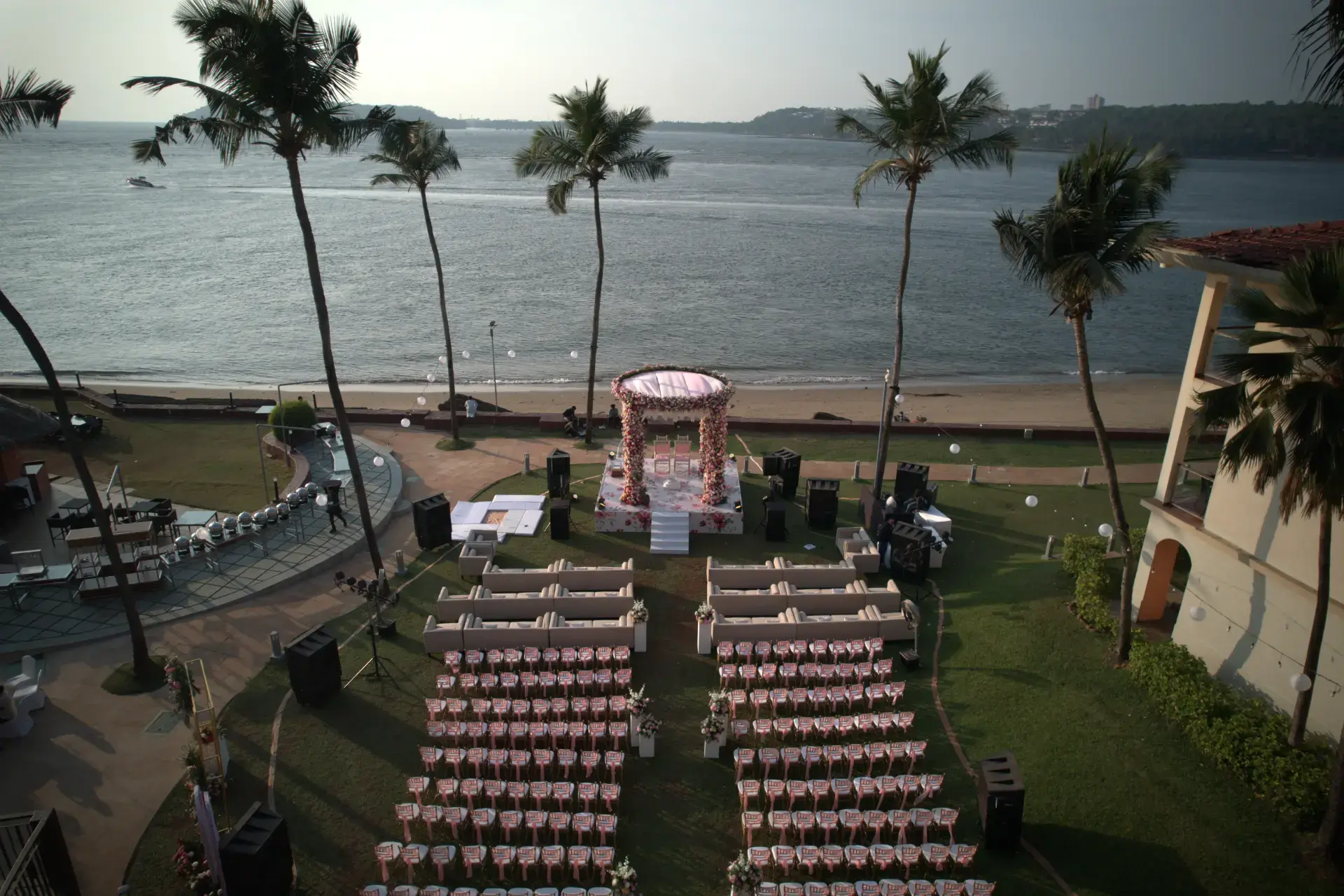 Aerial view of a beachfront wedding ceremony setup with a floral mandap facing the sea, rows of pink-and-white seating and palm trees along the coastline